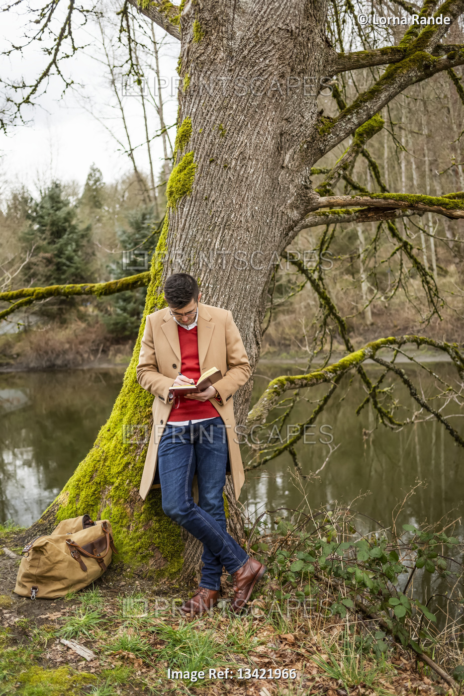 Young man in business casual attire leaning on a mossy tree beside a pond ...