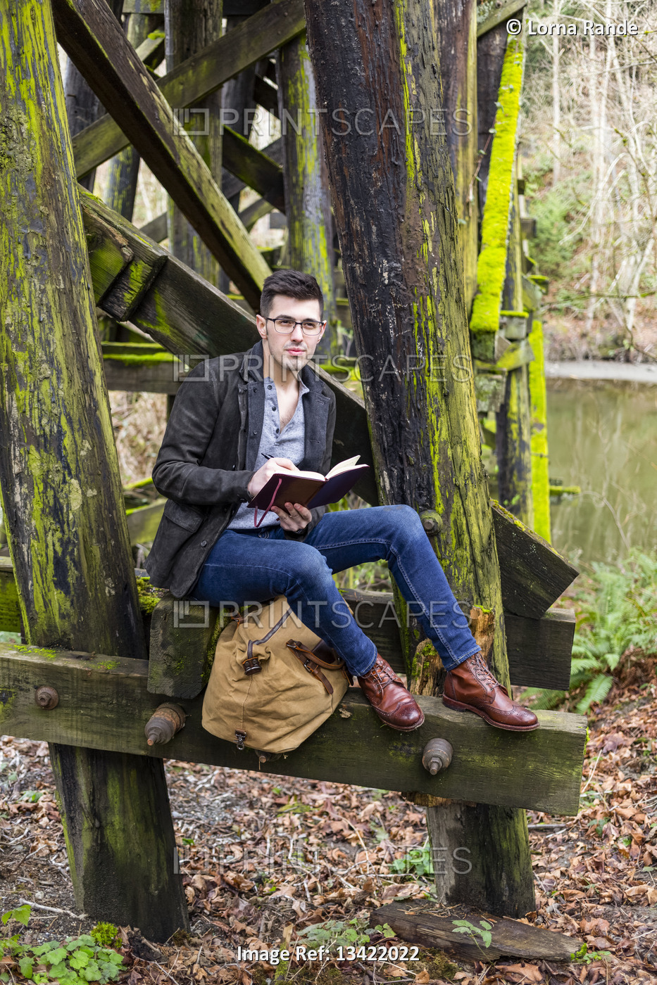 Young man in sitting on a moss-covered trestle bridge beside a pond reading ...