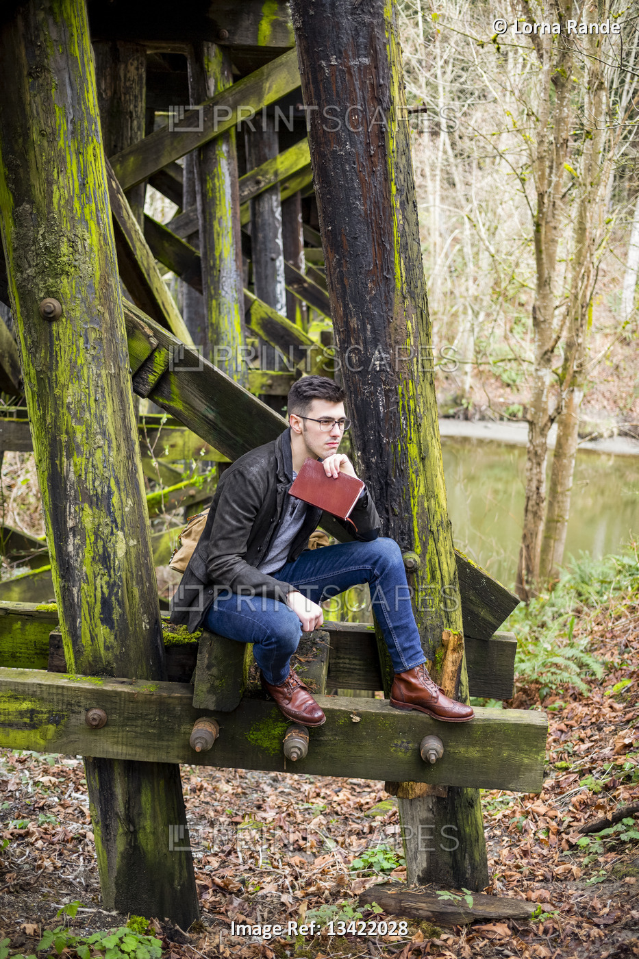 Young man in sitting on a moss-covered trestle bridge beside a pond with a book ...
