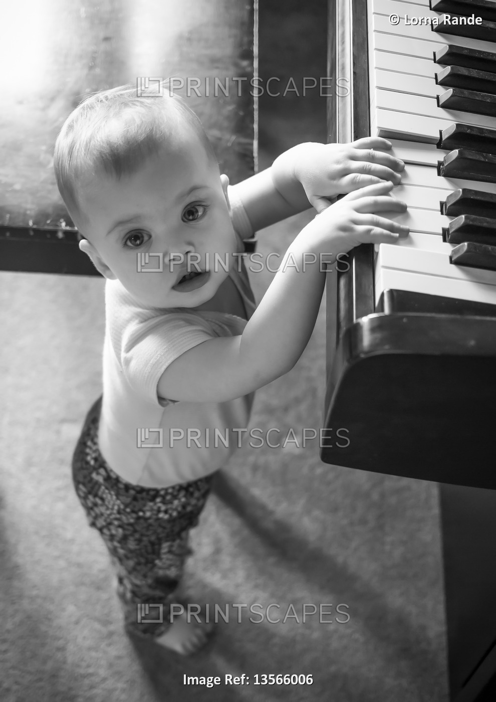 A baby girl standing and playing the piano while looking up at the camera; ...