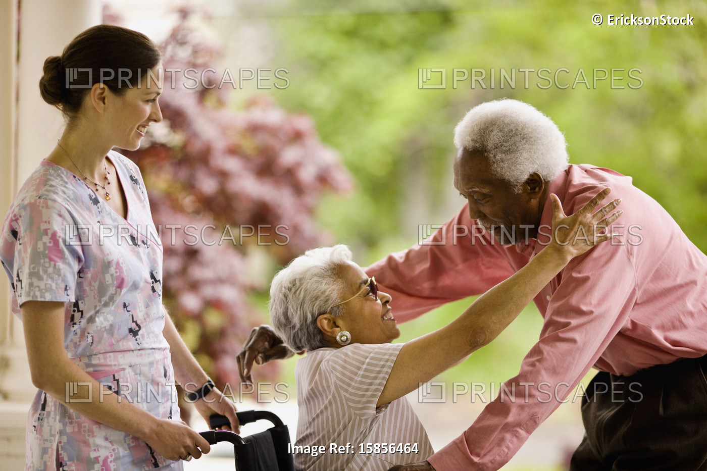 Nurse looks on as senior woman in wheelchair reaches up to embrace senior man ...