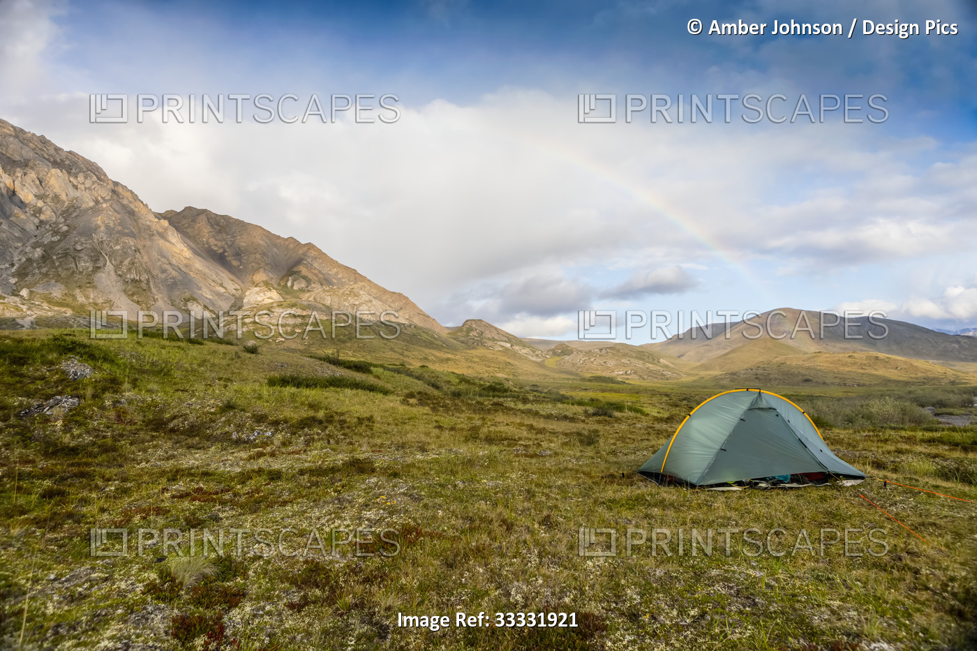 A rainbow arcs over a green backpacking tent set up on the tundra, mountains in ...