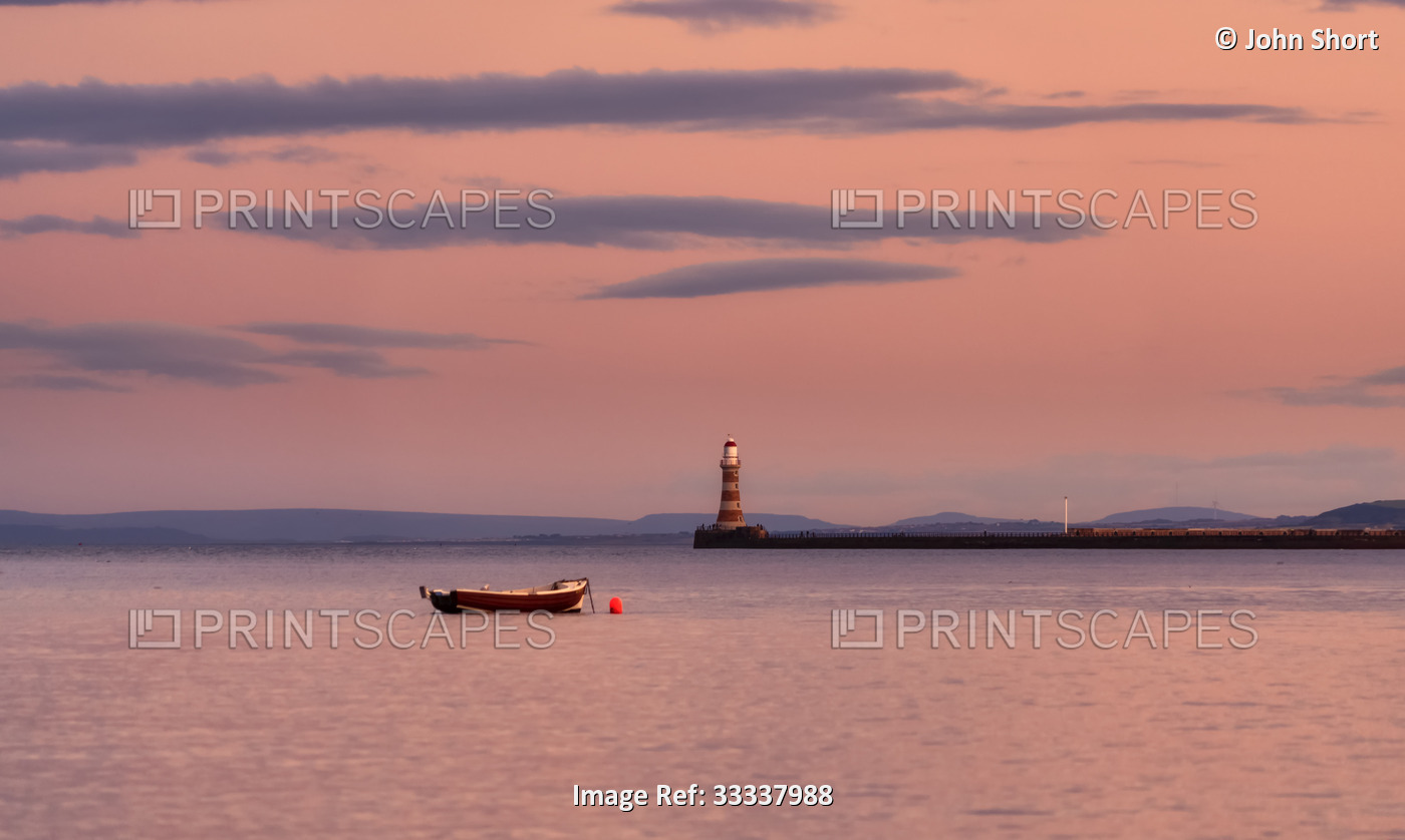 Roker Pier Lighthouse at dusk with a glowing pink sky reflected on the tranquil ...