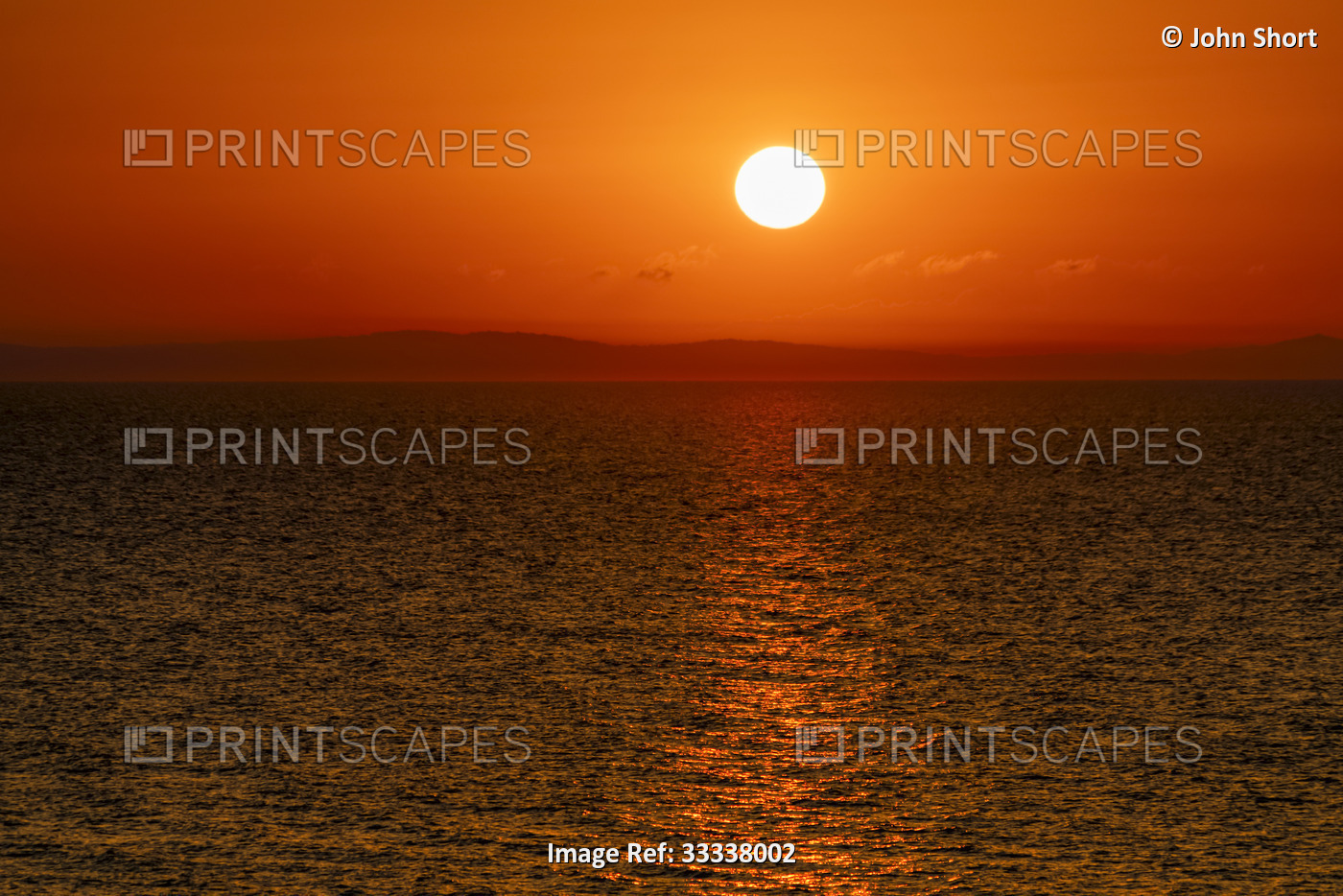Bright sun in a red sky at sunset over water and a silhouetted coastline; ...