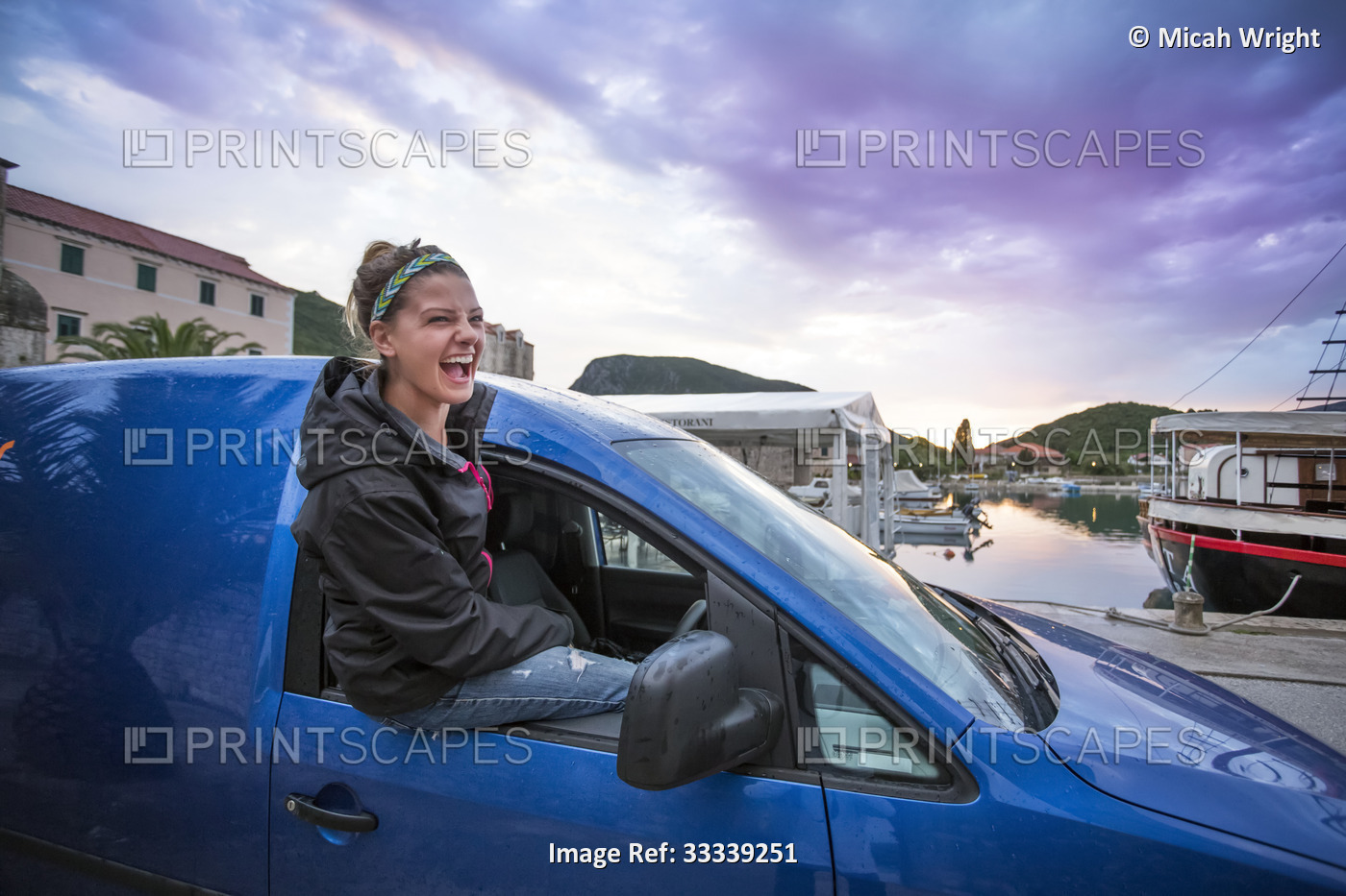 A young woman sits in the window of a car in a harbour at dusk. The lakeside ...