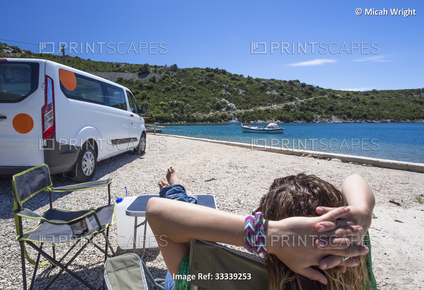 Woman sits in the sun resting outside of a camper van as group of travelers ...