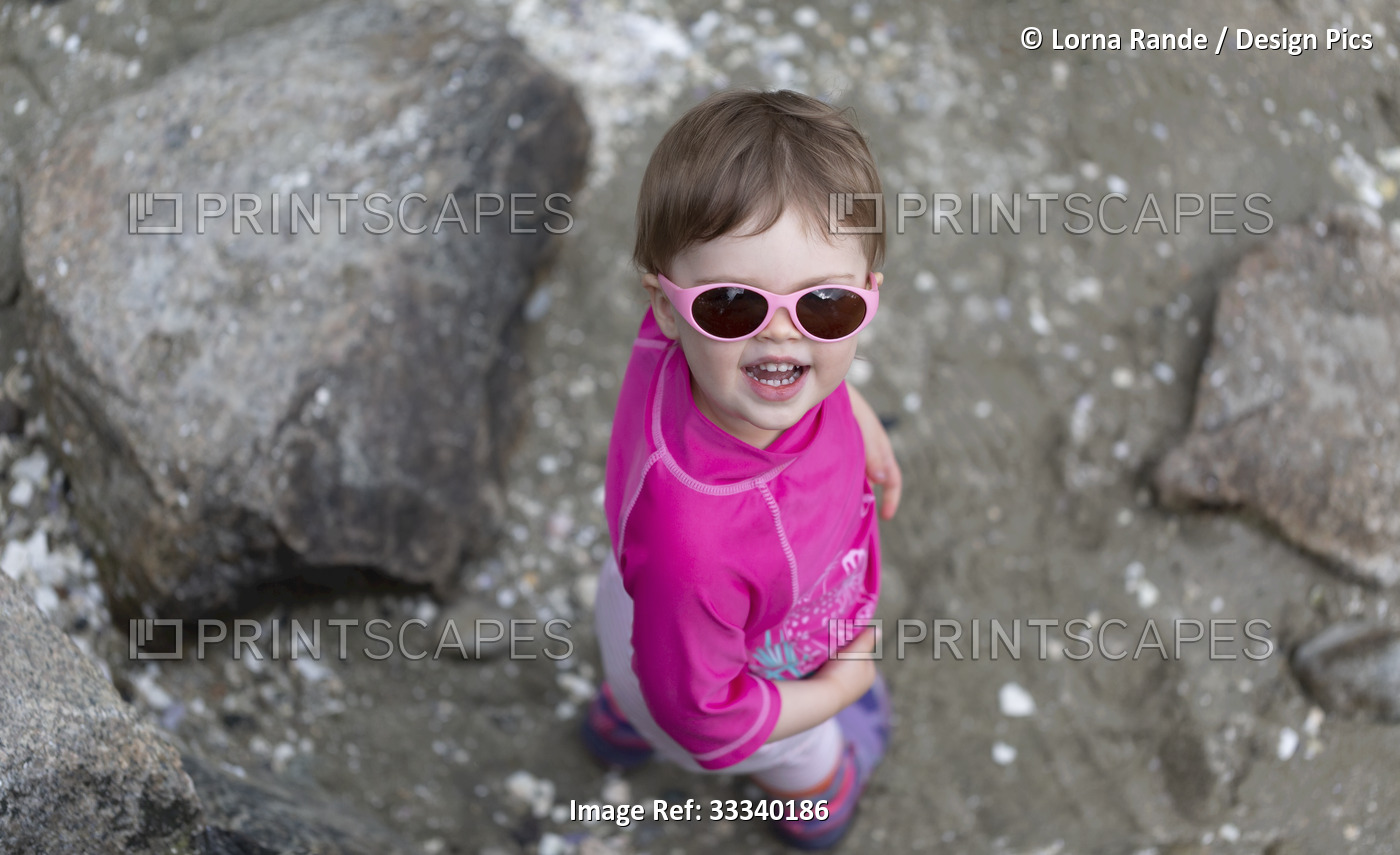 Portrait of 23 month old girl standing on a rocky beach wearing pink sunglasses ...