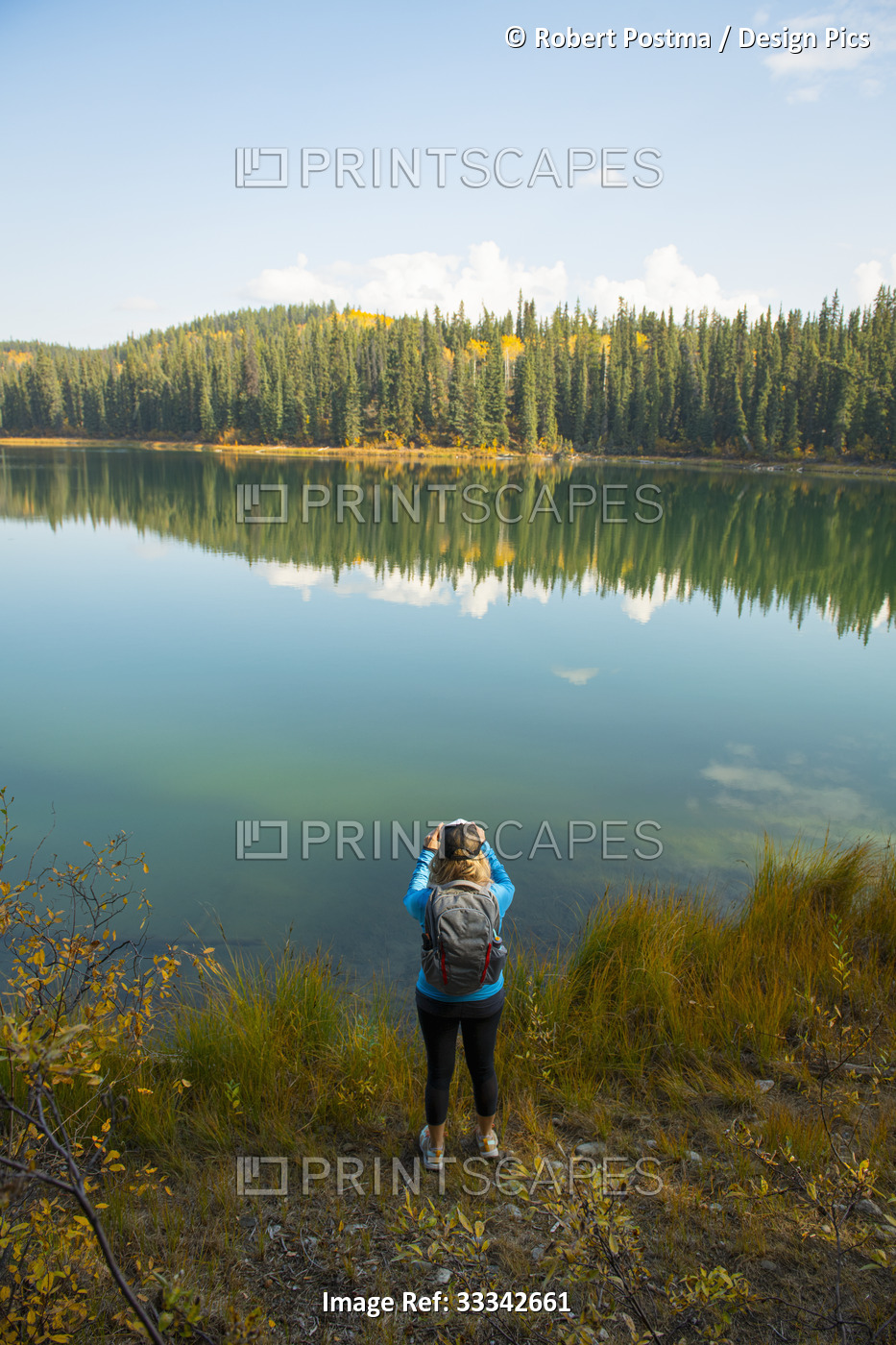 A woman stands at the water's edge looking out over a tranquil lake and taking ...