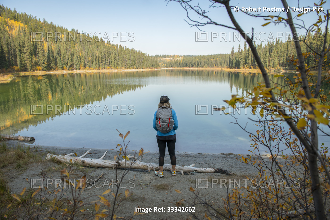 A woman stands at the water's edge looking out over a tranquil lake and taking ...