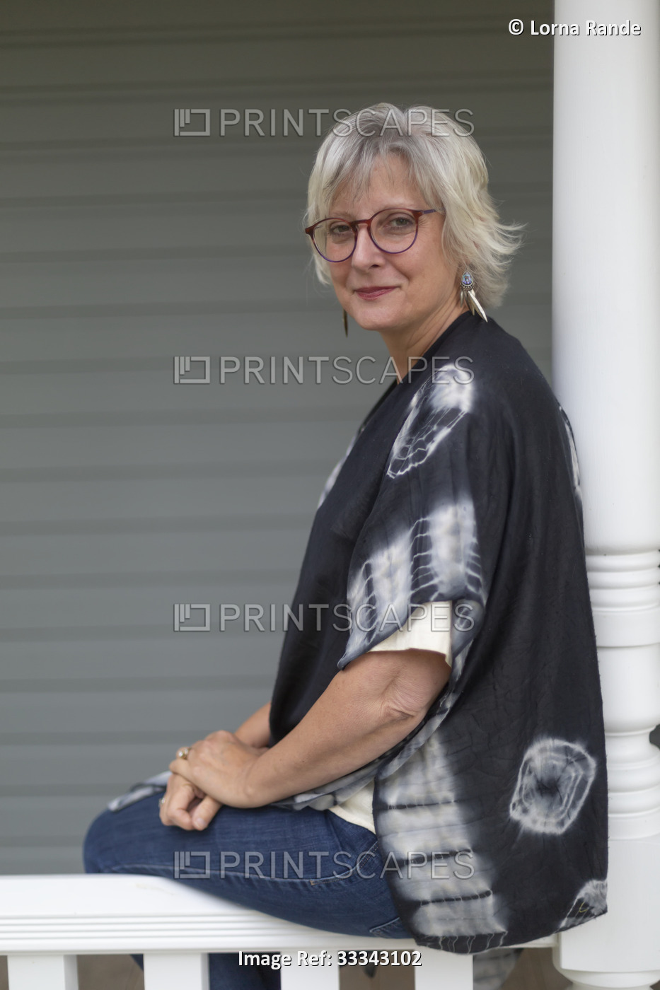 Portrait of a mature woman sitting on a porch railing in casual clothing; ...