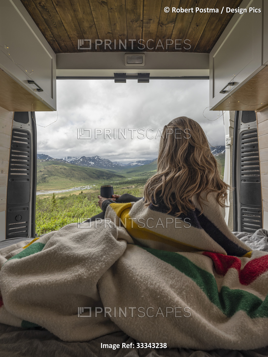 A woman sits under a Hudson's Bay point blanket in the back of a camper van and ...