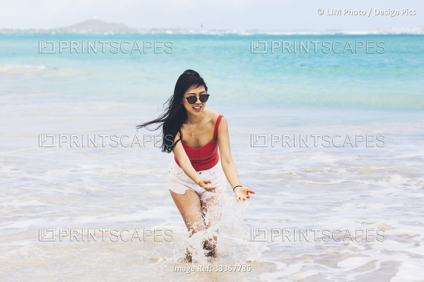 A woman stands in the surf, bending to touch the splashing water, as she enjoys ...