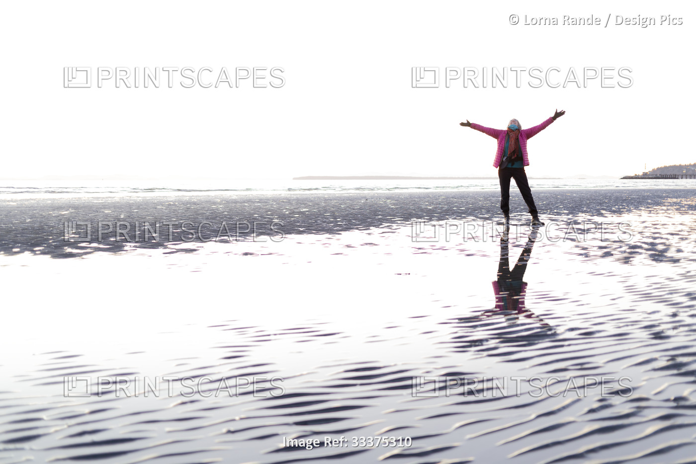 A senior woman stands on a wet beach with arms outstretched looking up to the ...