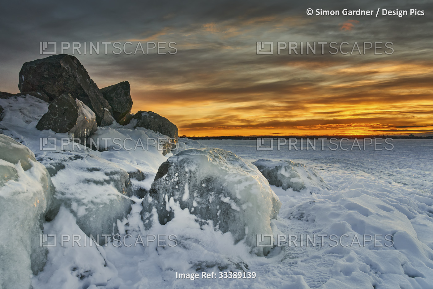 Rugged shoreline with ice-covered rocks and snow and golden glowing skies. This ...