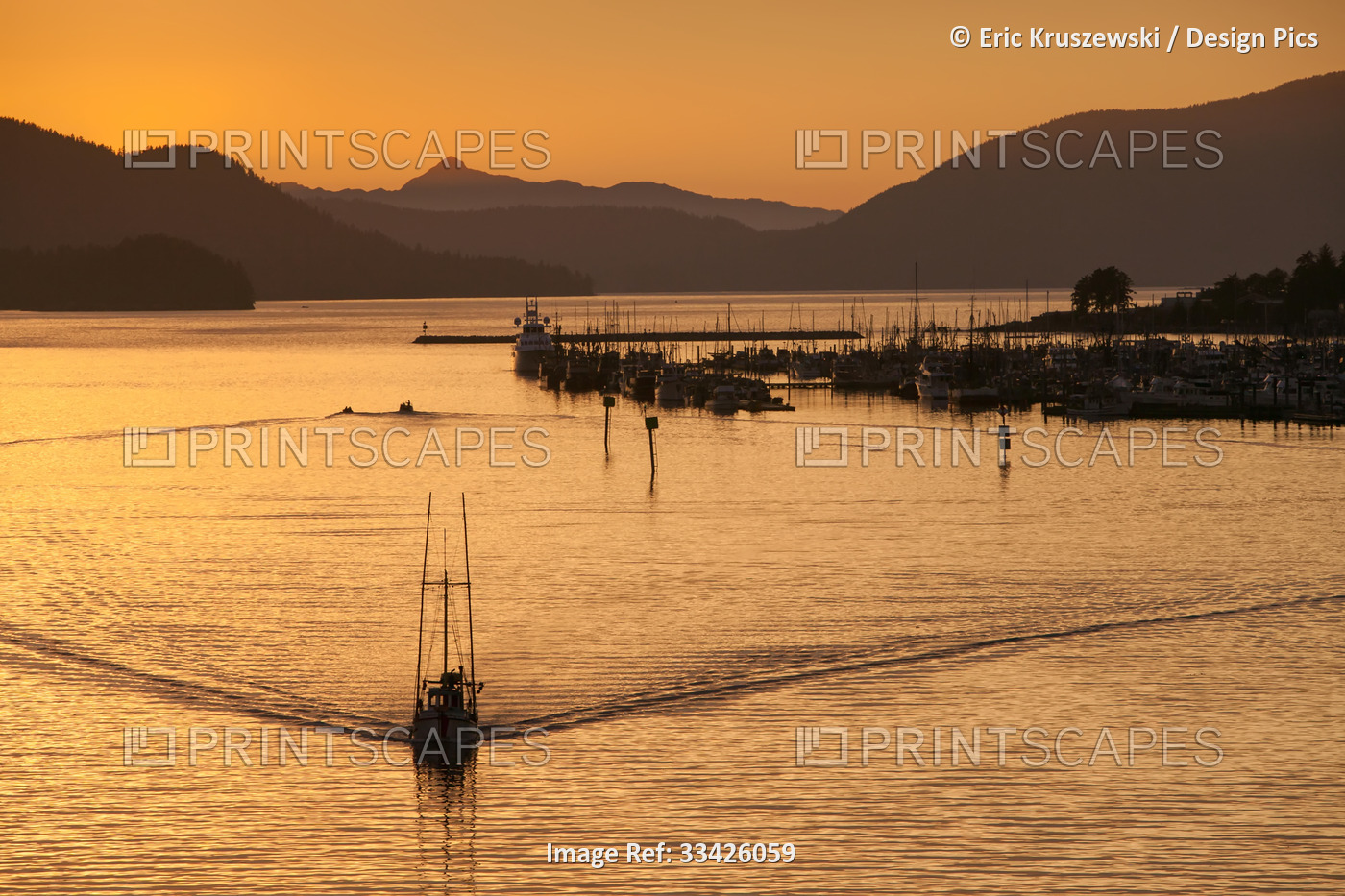 At sunset, a boat carves the water near mountains, a marina and harbor.; Sitka, ...