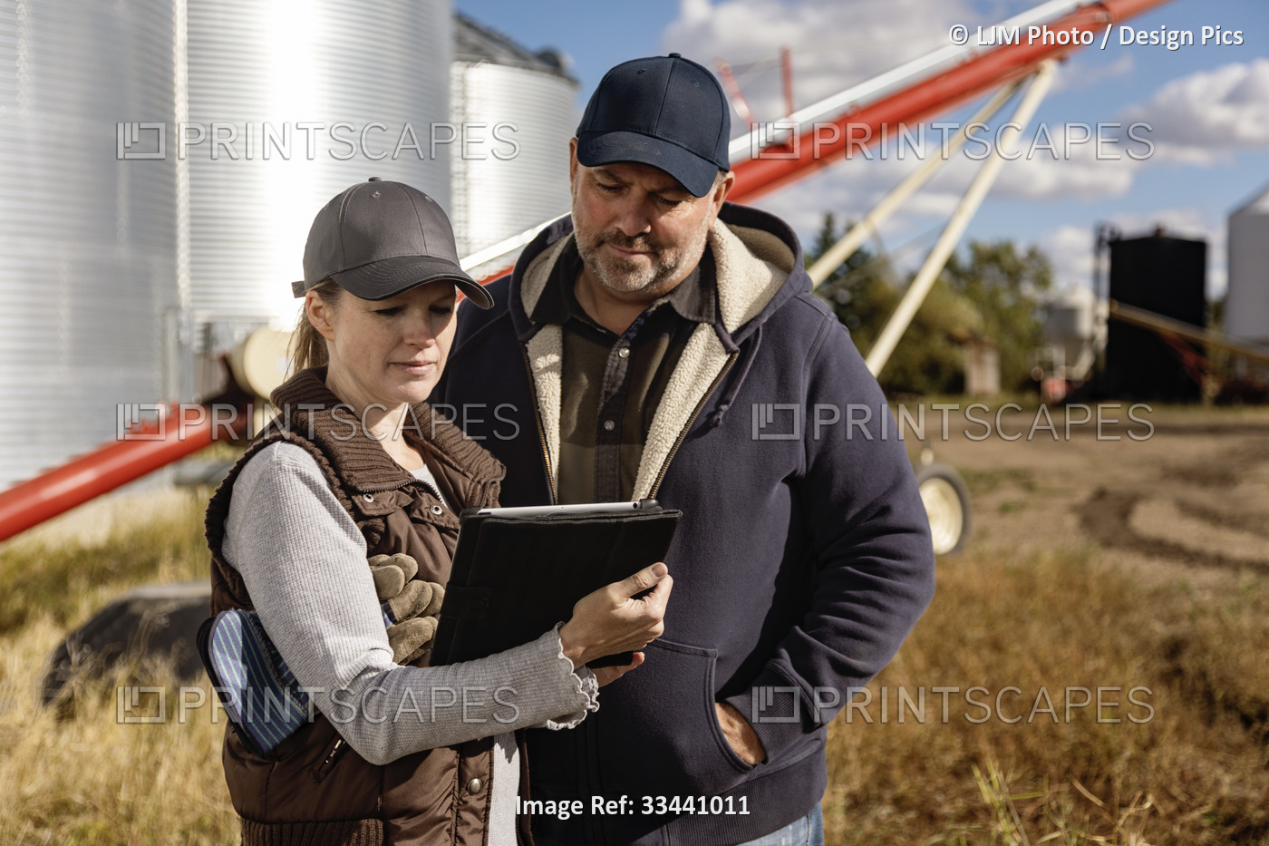 Mature couple working on their farm, standing in front of grain bins and auger ...