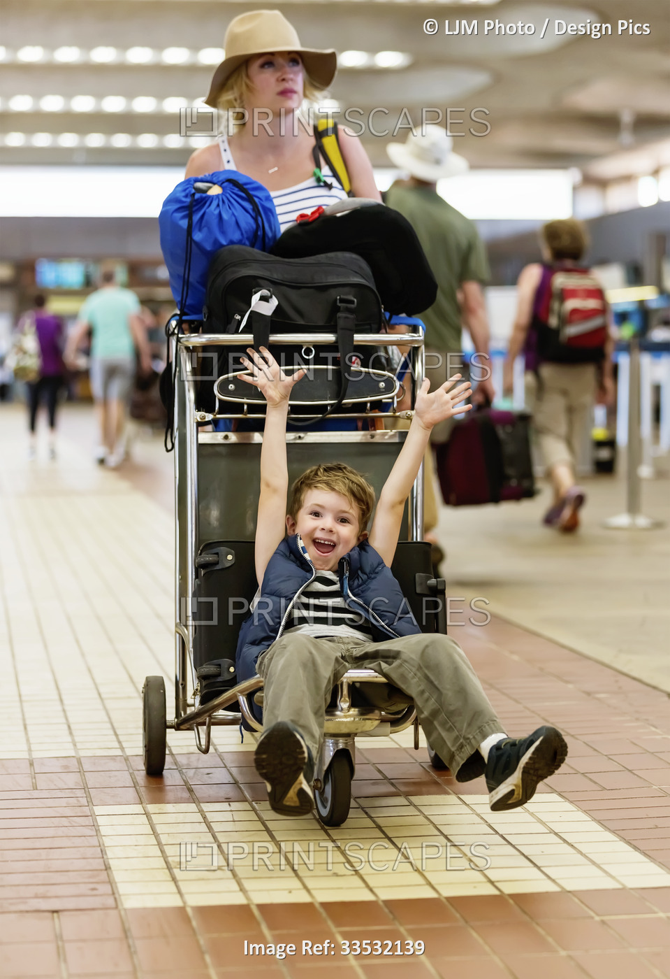 A mother pushes a luggage cart through an airport while her young son sits on ...