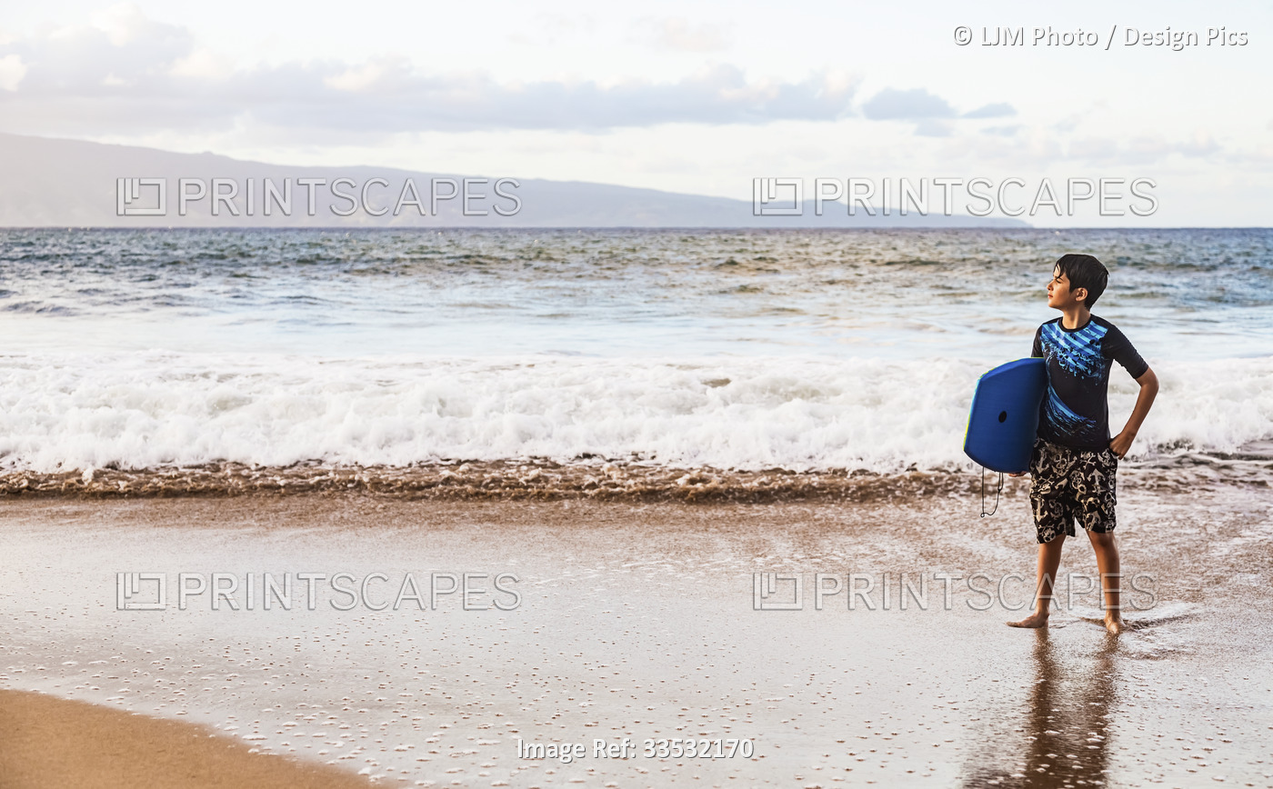 A boy stands on D. T. Fleming Beach at the water's edge holding a bodyboard ...