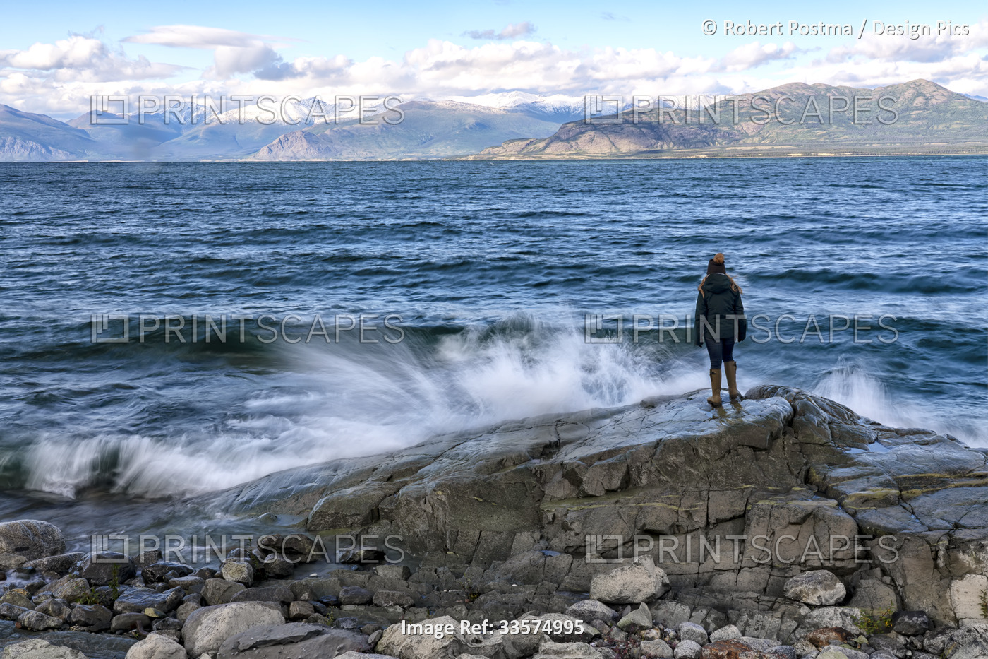 View taken from behind of a woman standing on a rock while the waves crah into ...