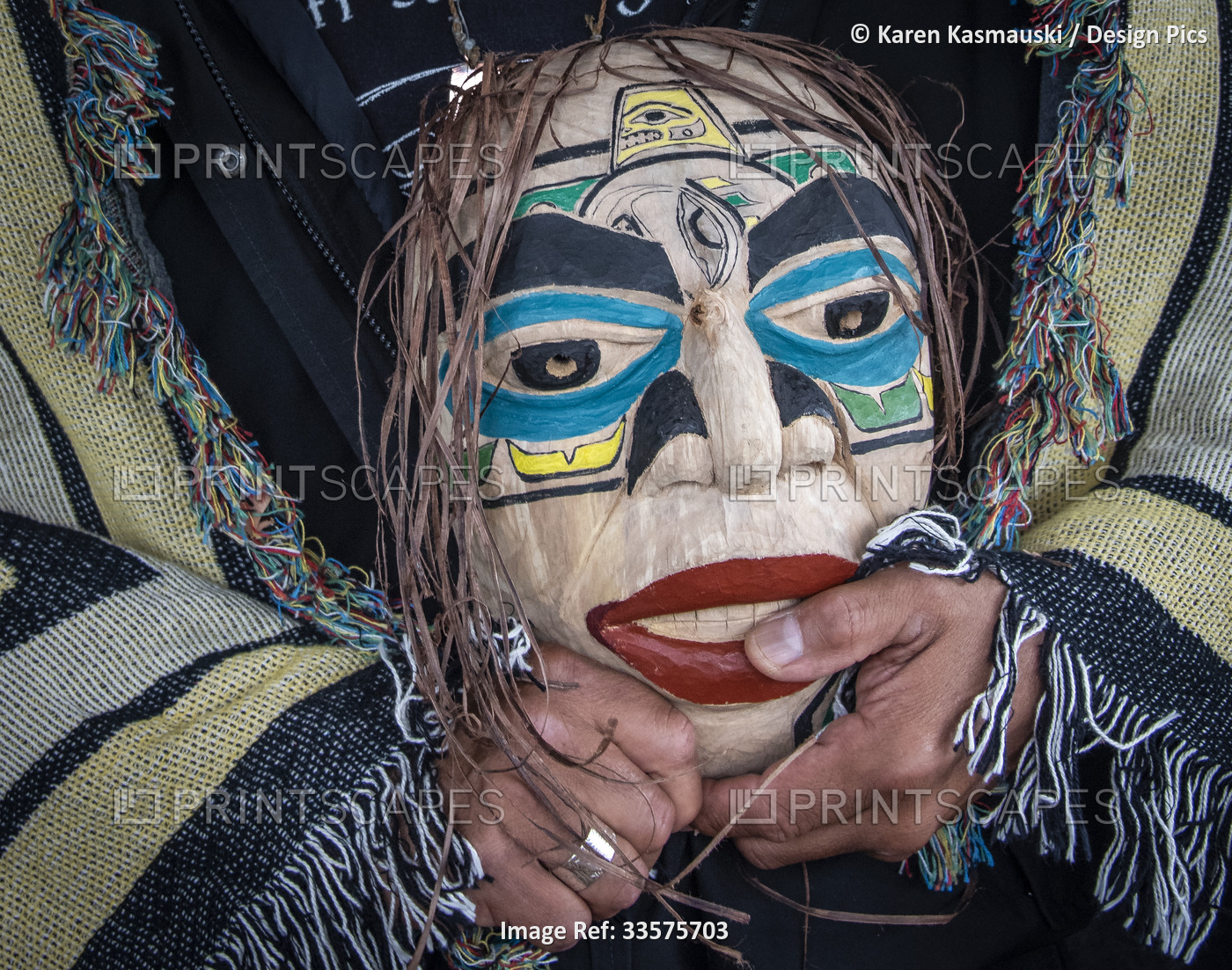Man stands draped in a blanket with Native Indian design and holding a mask; ...