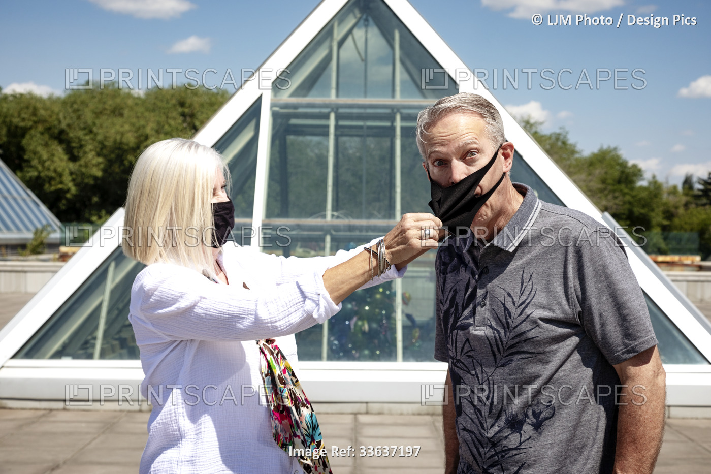 A mature couple stands outside wearing their face masks, the wife pulling the ...