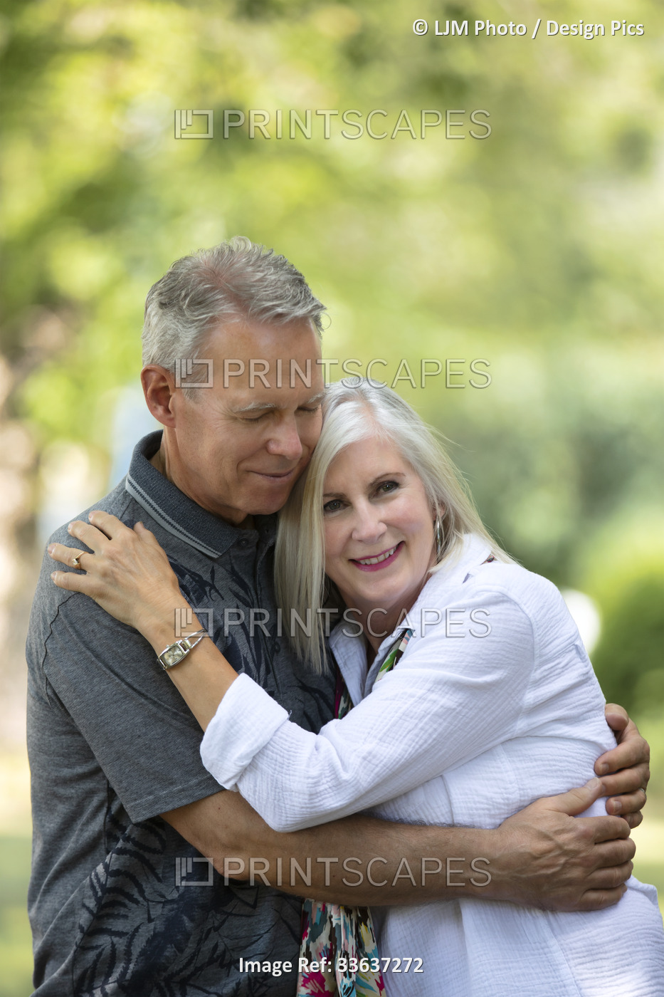 Mature couple sharing an embrace while walking outdoors on a park trail; ...
