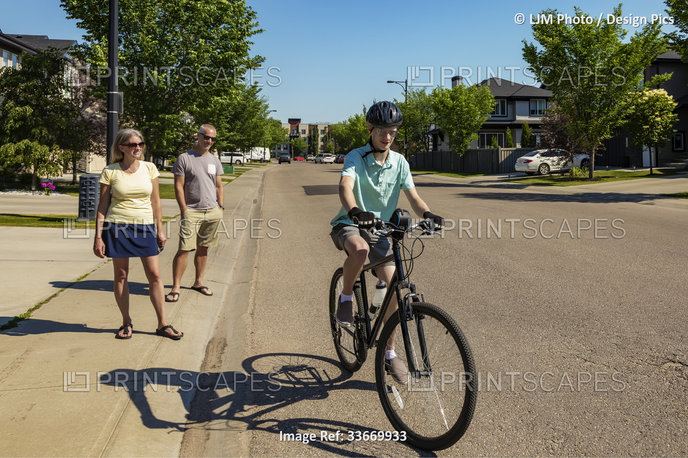 Young man riding his bike down a residential street, his parents standing on ...