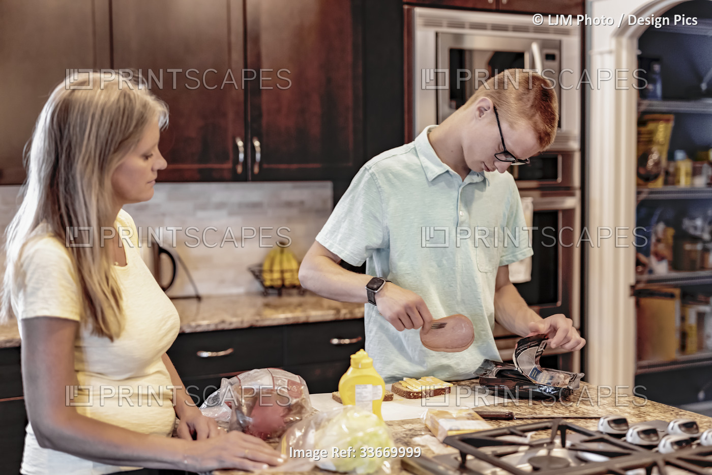 Young man prepares lunch in the kitchen at home as his mother looks on and ...