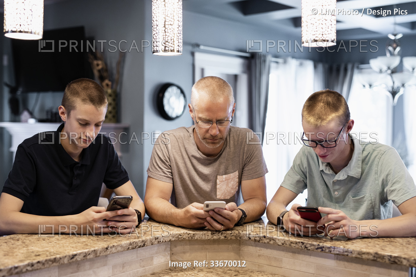Father and two sons sit at the kitchen island at home using their smart phones; ...