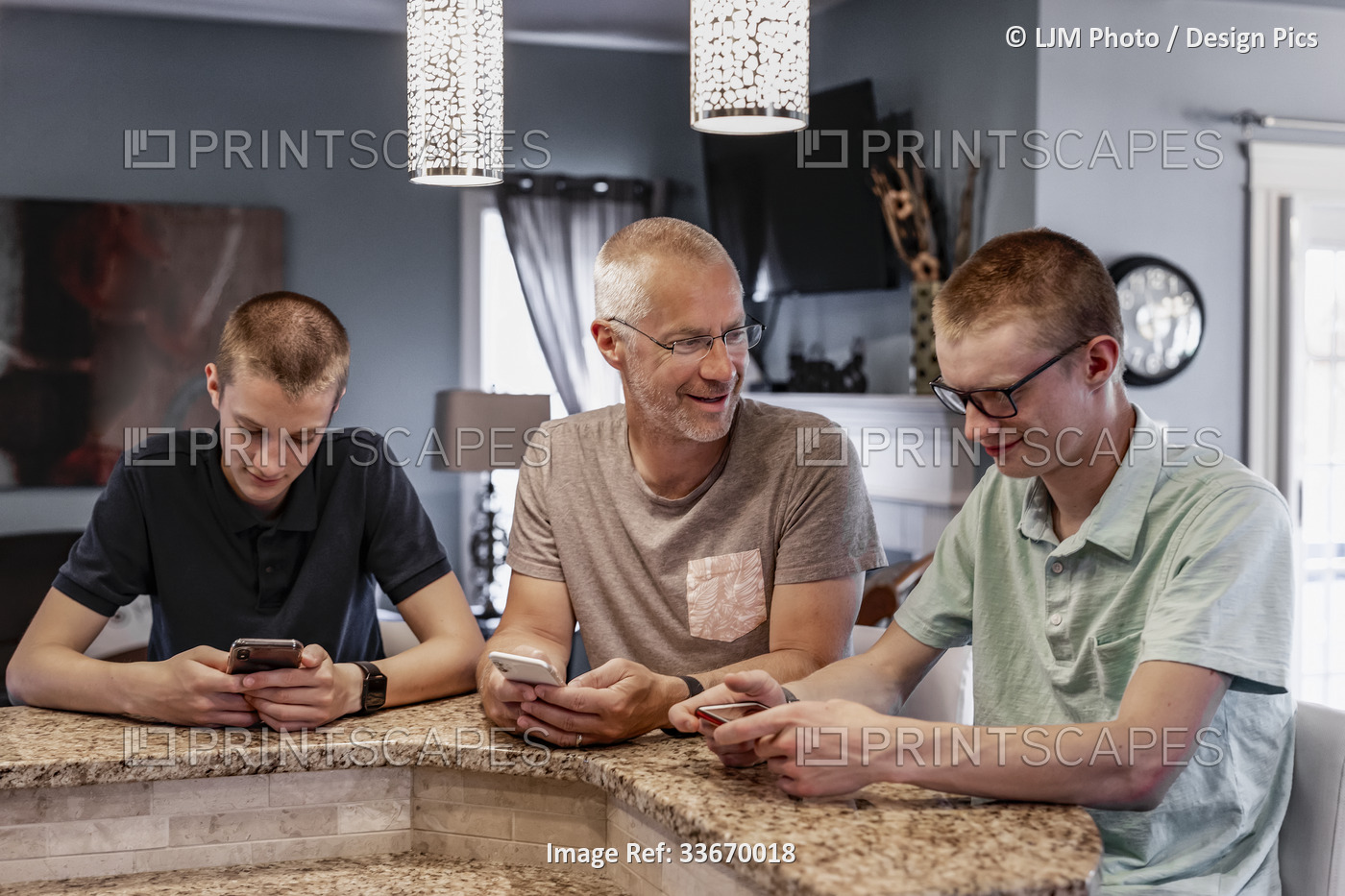 Father and two sons sit at the kitchen island at home using their smart phones, ...