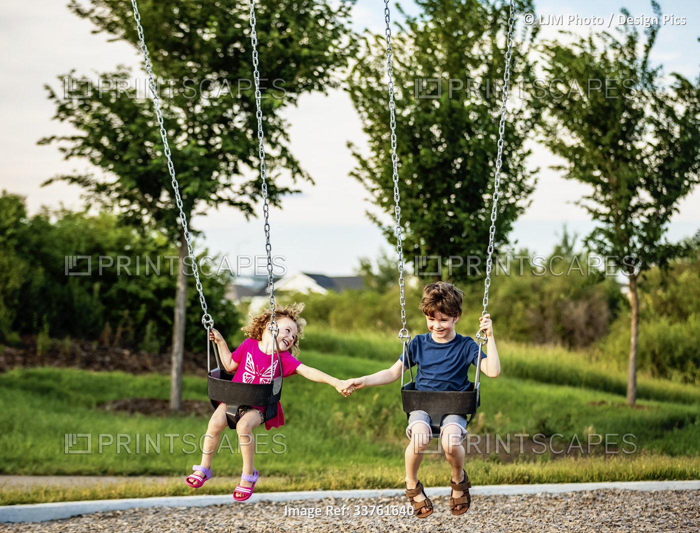 Young girl and boy holding hands while playing on a swing set at a playground; ...